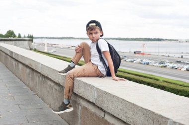 Beyaz tişörtlü ve sırt çantalı Avrupalı bir çocuk parkta oturuyor ve ileriye bakıyor.