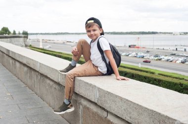 Beyaz tişörtlü ve sırt çantalı Avrupalı bir çocuk parkta oturuyor ve gülümsüyor.