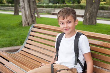 Beyaz tişörtlü ve sırt çantalı Avrupalı bir çocuk parkta bir bankta oturuyor ve gülümsüyor.