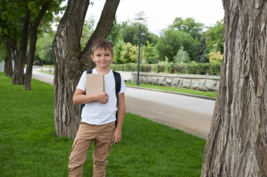 Beyaz tişörtlü, elinde kitap olan bir çocuk parkta yürüyor ve gülüyor. Okula geri dön.