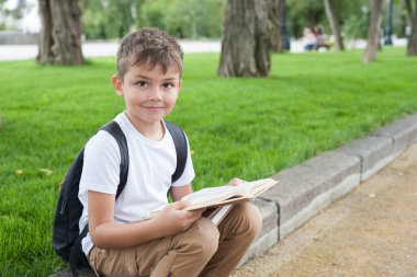 Beyaz tişörtlü, elinde kitap olan bir çocuk parkta yürüyor ve gülüyor. Okula geri dön.