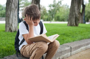 Beyaz tişörtlü, sırt çantalı Avrupalı bir çocuk parkta hüzünlü bir bakışla kitap okuyor.