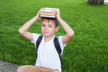 Beyaz tişörtlü, sırt çantalı bir çocuk kafasında bir yığın kitapla oturuyor. Okula geri dön.