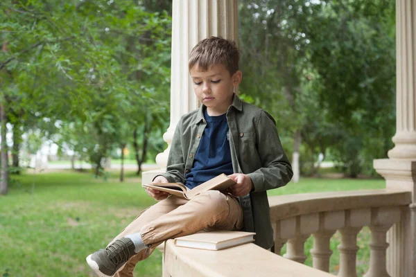 Avrupalı görünümlü bir çocuk parkta oturan bir kitabı dikkatle okur.