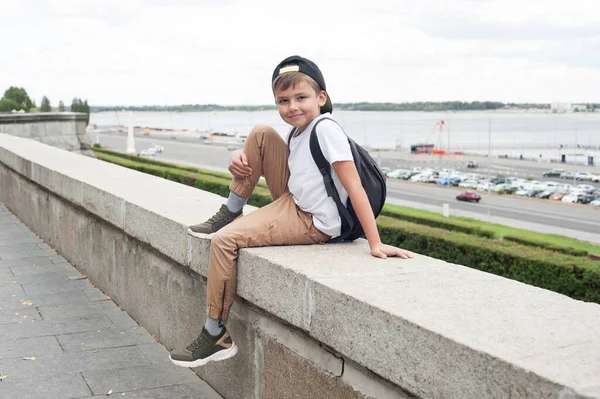 Beyaz tişörtlü ve sırt çantalı Avrupalı bir çocuk parkta oturuyor ve gülümsüyor.