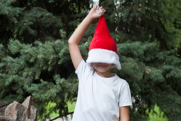 A boy in a white t shirt and Santa hat stands with the hat pulled over his face