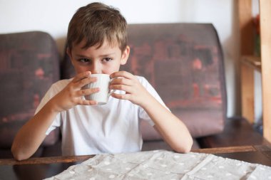 Avrupalı görünümlü bir çocuk evdeki kanepede oturan beyaz bardaktan içer.