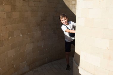 Beyaz tişörtlü Avrupalı bir çocuk duvarın arkasından gülümseyerek dışarı bakar. Açık hava oyunları. Saklambaç