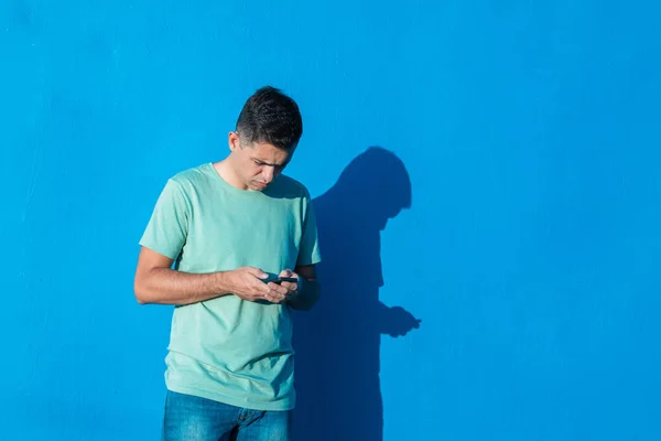 Młody człowiek za pomocą swojego smartfona surfowanie po Internecie stoi izolowane na niebieskim tle. Koncepcja ludzi i emocji z przestrzenią kopiowania. — Zdjęcie stockowe