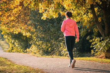 Sonbahar ormanlarında gün batımında koşan ve pembe ceketli bir kız. Sonbahar renklerinde koşan genç bir kadın. Spor ve aktivite konsepti.