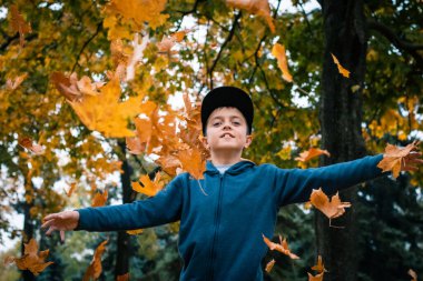 Mavi kapüşonlu ve siyah kapüşonlu bir çocuk sonbahar parkında uçan sarı, turuncu ve kırmızı yaprakların arasında atlıyor. Renkli orman ve mevsimlik park kavramı.