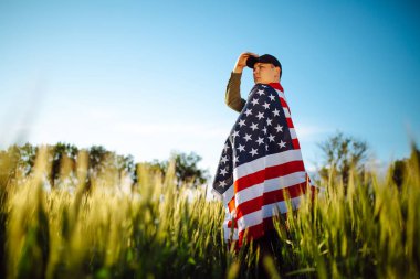 Yeşil buğday tarlasında Amerikan bayrağına sarılı yeşil gömlek ve şapka giyen genç bir adam. Vatansever çocuk 4 Temmuz 'da elinde ulusal bayrağıyla bağımsızlık gününü kutluyor.