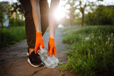 Genç adam gönüllü olarak parktan çöp topluyor. Plastik şişe turuncu eldiven giyen adam tarafından toplanıyor. Kirlilik, ekoloji, çevre kavramı