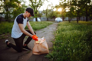 Genç adam gönüllü olarak parktan çöp topluyor. Plastik şişe turuncu eldiven giyen adam tarafından toplanıyor. Kirlilik, ekoloji, çevre kavramı
