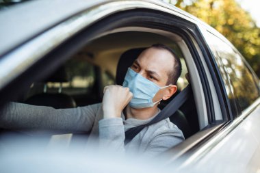 Bir adam araba kullanıyor ve Coronavirus sırasında tıbbi maskeye öksürüyor. Taksi şoförü, yolcularını gideceği yere götürür. Sosyal mesafe, yeni normal, virüs yayılmış önleme ve tedavi kavramı