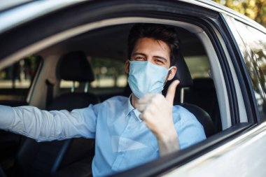 Genç taksi şoförü, arabada oturan ve koruyucu steril tıbbi maske takan, koronavirüs salgını sırasında çok çalışan bir baş parmak işareti gösteriyor. Sosyal mesafe, virüs yayılması önleme, tedavi konsepti