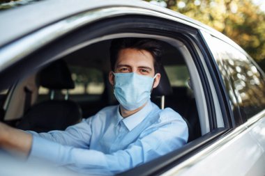 Genç adam steril tıbbi maske takarak direksiyon başında oturuyor. Erkek taksi şoförü Coronavirus salgını sırasında çok çalışıyor. Sosyal mesafe, virüs yayılımı önleme ve tedavi kavramı