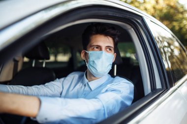 Genç adam steril tıbbi maske takarak direksiyon başında oturuyor. Erkek taksi şoförü Coronavirus salgını sırasında çok çalışıyor. Sosyal mesafe, virüs yayılımı önleme ve tedavi kavramı