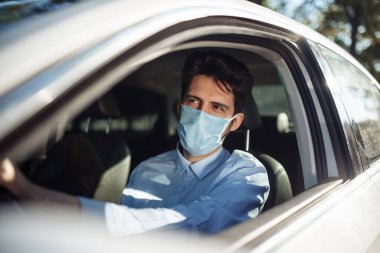 Genç adam steril tıbbi maske takarak direksiyon başında oturuyor. Erkek taksi şoförü Coronavirus salgını sırasında çok çalışıyor. Sosyal mesafe, virüs yayılımı önleme ve tedavi kavramı