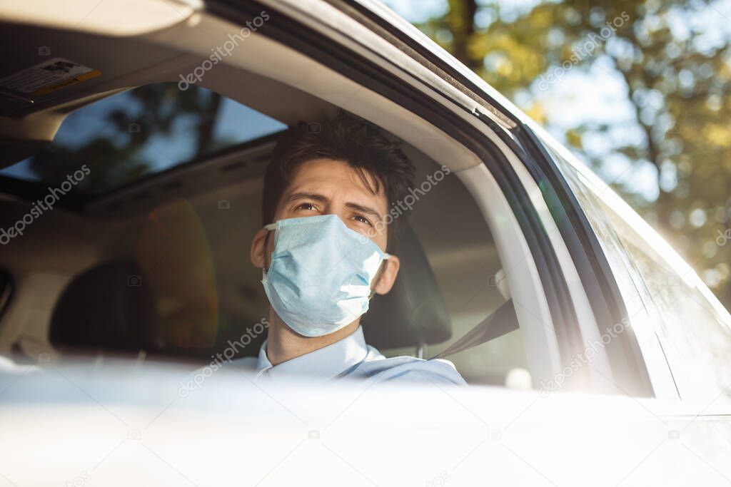 Young man sits behind the steering wheel in the car wearing sterile medical mask. Boy taxi driver works hard during coronavirus outbreak. Social distance, virus spread prevention and treat concept