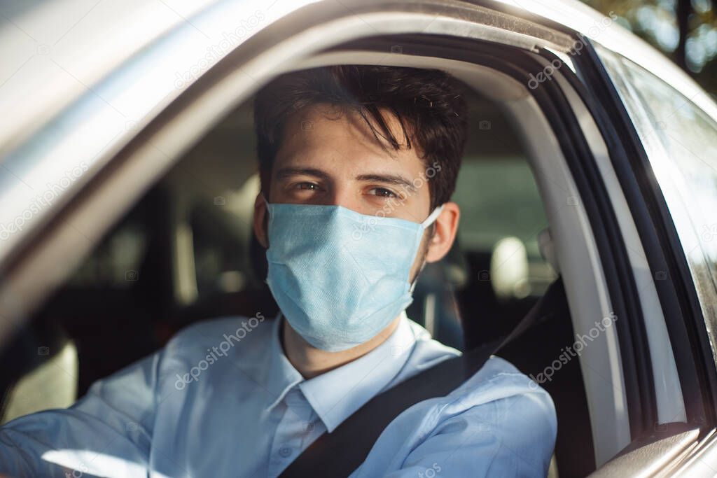 Closeup portrait of young man sits in the car wearing sterile medical mask. Boy taxi driver works hard during coronavirus outbreak. Social distance, virus spread prevention and treat concept
