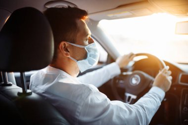 Genç taksi şoförü Coronavirus salgını sırasında steril tıbbi maske takıyor. Bir çocuk gün batımında trafikte bir araba sürer. Sosyal mesafe ve sağlık hizmeti kavramı