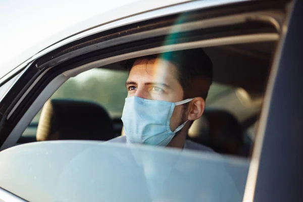 Young businessman takes a taxi and looks out of the car window wearing sterile medical mask. A man sits on the back seat of taxi and takes a ride during coronavirus pandemic. Social distance concept