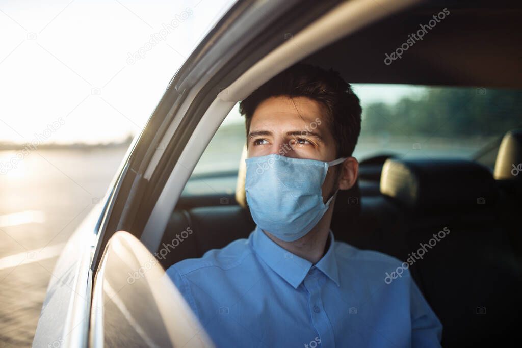 Young businessman takes a taxi and looks out of the car window wearing sterile medical mask. A man sits on the back seat of taxi and takes a ride during coronavirus pandemic. Social distance concept