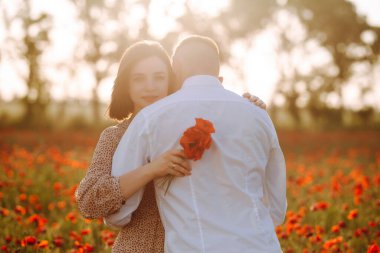 Günbatımında kırmızı gelincik tarlasında kucaklaşan romantik çift. Oğlan ve kız birbirlerine sevdiklerini gösteriyorlar ellerinde kırmızı gelincik çiçeği buketi var. Aşk, aile değerleri ve barış kavramı
