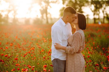 Genç ve romantik evli çift, güzel günbatımında kırmızı gelincik çiçeklerinin arasında duruyor. Çekici erkek ve hassas kız mutlu hissederek sarılıp öpüşürler. Aşk, aile kavramı