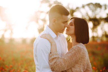 Genç ve romantik evli çift, güzel günbatımında kırmızı gelincik çiçeklerinin arasında duruyor. Çekici erkek ve hassas kız mutlu hissederek sarılıp öpüşürler. Aşk, aile kavramı