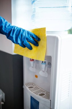 Coronavirus pandemik karantinası sırasında iş yerinde mavi lastik eldiven giyip sarı bez ile su soğutucuyu temizleyen bir el. Sağlık ve adaptasyon kavramı