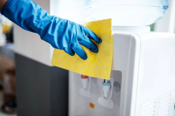 Coronavirus pandemik karantinası sırasında iş yerinde mavi lastik eldiven giyip sarı bez ile su soğutucuyu temizleyen bir el. Sağlık ve adaptasyon kavramı
