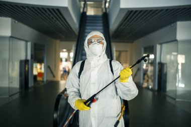 Boş bir alışveriş merkezindeki yürüyen merdivenin yanında dezenfekte elbisesi giymiş bir adamın portresi. Covid-19 'u önlemek için halka açık yerleri temizleyen bir gönüllü. Sağlık farkındalığı kavramı