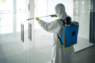 Dezenfekte elbisesi giymiş bir adam, cam kapı kollarına dezenfektan sıkmış. Boş bir alışveriş merkezinde, kovid-19 yayılmasını önlemek için. Sağlık farkındalığı, temizlik, savunma kavramı