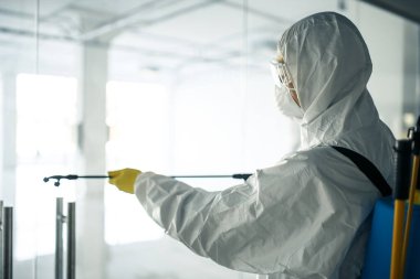 Dezenfekte elbisesi giymiş bir adam, cam kapı kollarına dezenfektan sıkmış. Boş bir alışveriş merkezinde, kovid-19 yayılmasını önlemek için. Sağlık farkındalığı, temizlik, savunma kavramı