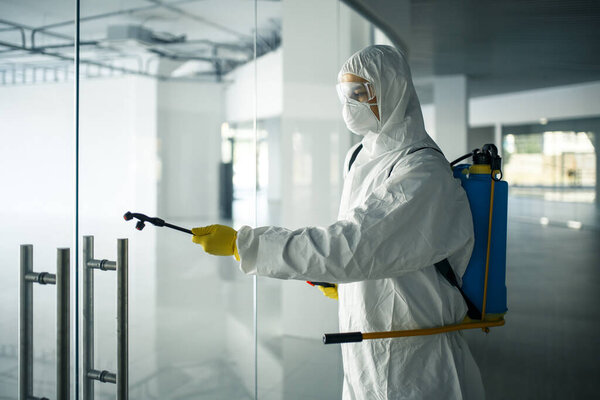A man wearing disinfection suit spraying with sanitizer the glass doors' handles in an empty shopping mall to prevent covid-19 spread. Health awareness, clean, defence concept