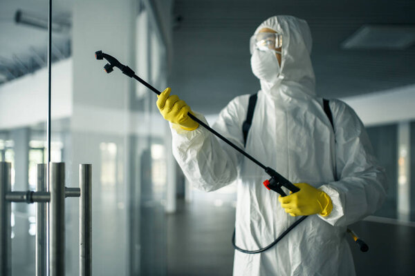 A man wearing disinfection suit spraying with sanitizer the glass doors' handles in an empty shopping mall to prevent covid-19 spread. Health awareness, clean, defence concept