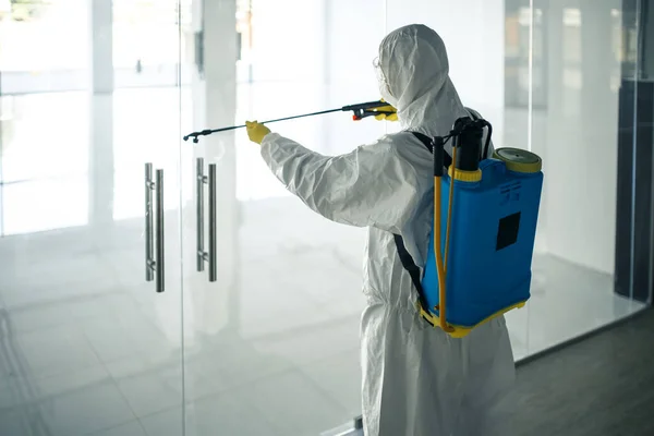 A man wearing disinfection suit spraying with sanitizer the glass doors\' handles in an empty shopping mall to prevent covid-19 spread. Health awareness, clean, defence concept