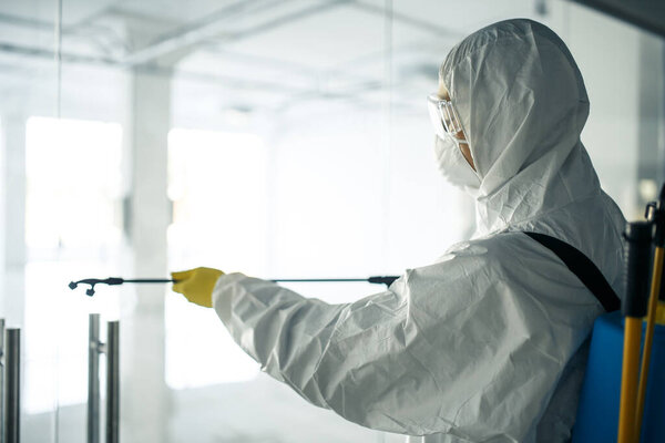 A man wearing disinfection suit spraying with sanitizer the glass doors' handles in an empty shopping mall to prevent covid-19 spread. Health awareness, clean, defence concept