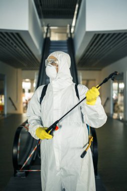 Boş bir alışveriş merkezindeki yürüyen merdivenin yanında dezenfekte elbisesi giymiş bir adamın portresi. Covid-19 'u önlemek için halka açık yerleri temizleyen bir gönüllü. Sağlık farkındalığı kavramı