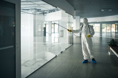 Dezenfekte elbisesi giymiş bir adam, cam kapı kollarına dezenfektan sıkmış. Boş bir alışveriş merkezinde, kovid-19 yayılmasını önlemek için. Sağlık farkındalığı, temizlik, savunma kavramı