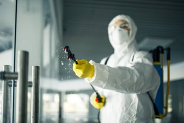 Dezenfekte elbisesi giymiş bir adam, cam kapı kollarına dezenfektan sıkmış. Boş bir alışveriş merkezinde, kovid-19 yayılmasını önlemek için. Sağlık farkındalığı, temizlik, savunma kavramı