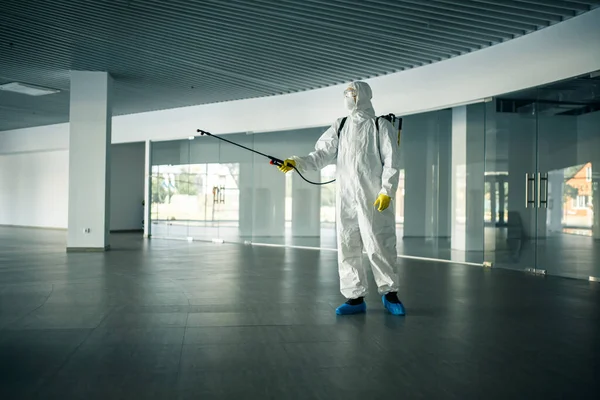 A man wearing disinfection suit spraying with sanitizer the glass doors\' handles in an empty shopping mall to prevent covid-19 spread. Health awareness, clean, defence concept