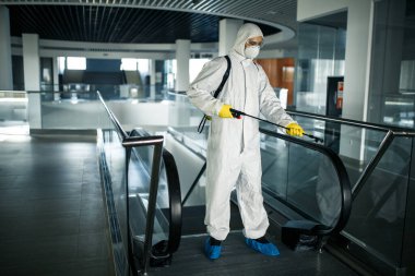 Koruyucu giysi giyen bir dezenfeksiyon işçisi boş bir iş merkezinde profesyonel olarak yürüyen merdiveni temizliyor. Antibakteriyel giysilerle donatılmış bir adam alışveriş merkezini sterilize eder. Kapsamlı kavram