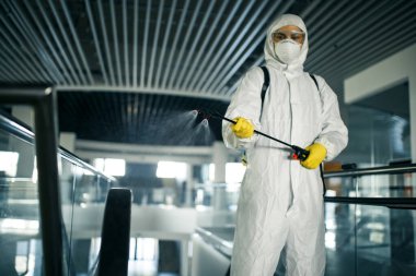 Koruyucu giysi giyen bir dezenfeksiyon işçisi boş bir iş merkezinde profesyonel olarak yürüyen merdiveni temizliyor. Antibakteriyel giysilerle donatılmış bir adam alışveriş merkezini sterilize eder. Kapsamlı kavram