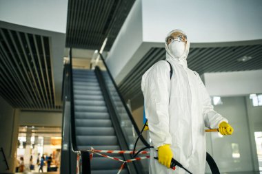 Boş bir alışveriş merkezindeki yürüyen merdivenin yanında dezenfekte elbisesi giymiş bir adamın portresi. Covid-19 'u önlemek için halka açık yerleri temizleyen bir gönüllü. Sağlık farkındalığı kavramı