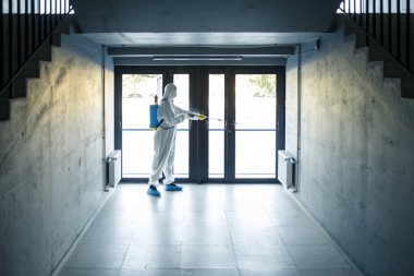 A man wearing protective disinfection suit and a spray stands in front of a glass doors under the staircases. Worker cleaning up the business center. Healthcare, covid-19 concept clipart