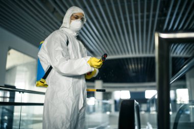 Koruyucu giysi giyen bir dezenfeksiyon işçisi boş bir iş merkezinde profesyonel olarak yürüyen merdiveni temizliyor. Antibakteriyel giysilerle donatılmış bir adam alışveriş merkezini sterilize eder. Kapsamlı kavram