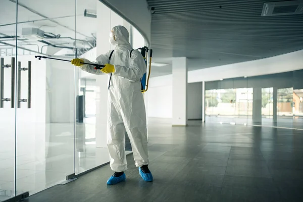 A man wearing disinfection suit spraying with sanitizer the glass doors\' handles in an empty shopping mall to prevent covid-19 spread. Health awareness, clean, defence concept